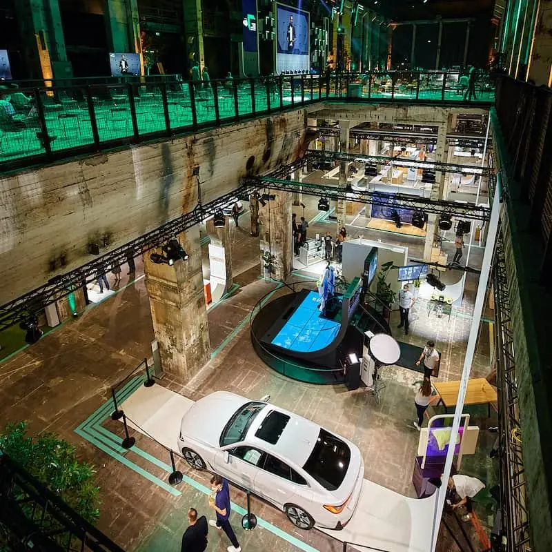 An exhibition hall in green light seen from the gallery above, visitors between the stands
