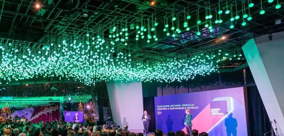 A full conference hall under hundreds of hanging green lights, a speaker on stage